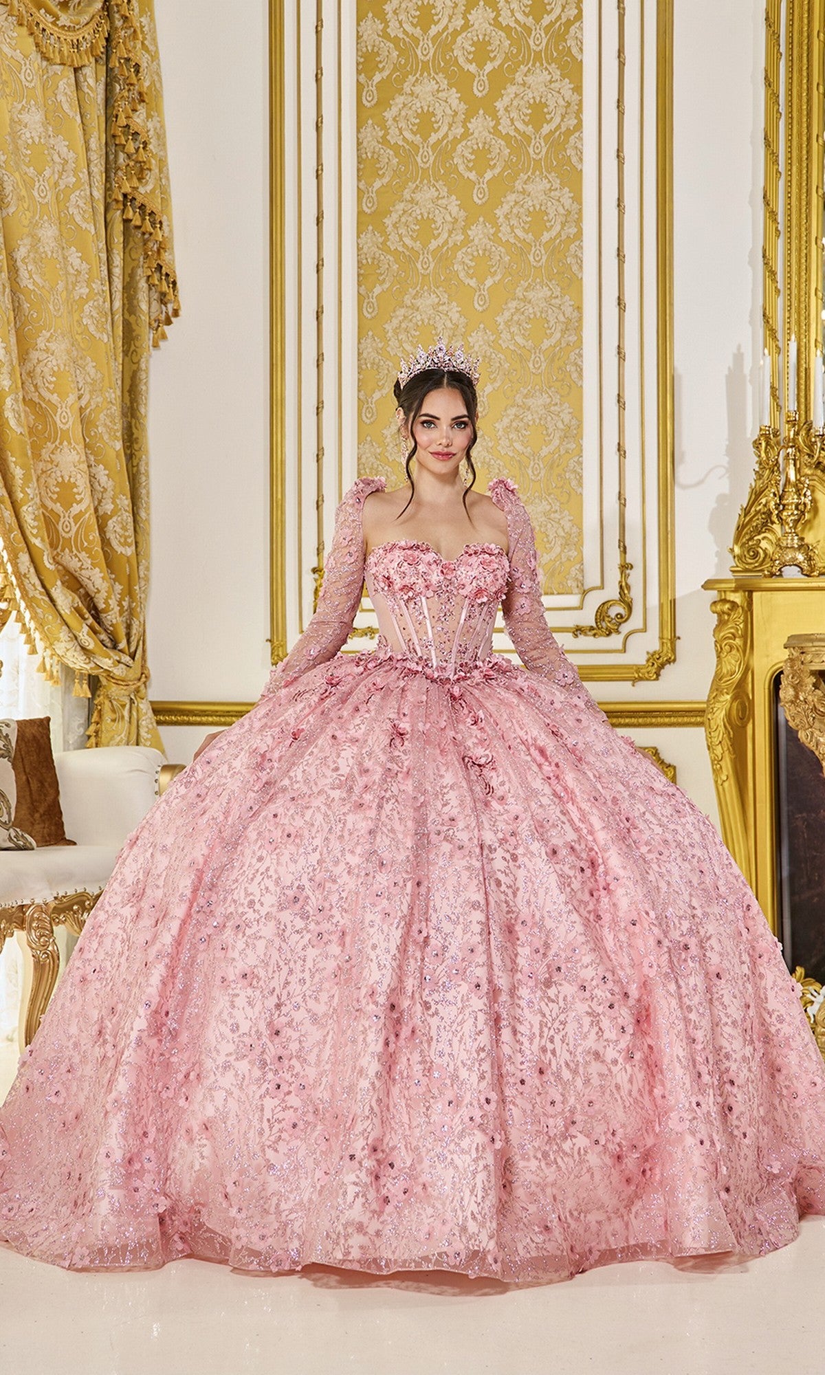 A young woman in the Quinceanera Dress 5077 By Dancing Queen, featuring a blush color and corset bodice, poses with a tiara in an elegant room decorated in gold and white.